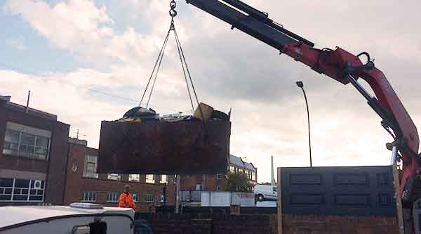 distance photo of a jcb arm with a large factory metal liquid container hanging from it being loaded on to a howarth scrap mans vehicle