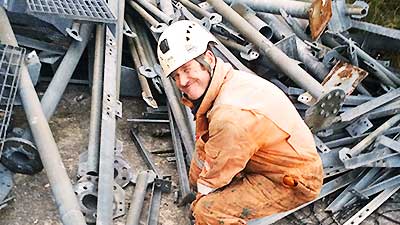close up of howarths scrap company sheffield manager dressed in the usual high visibilty gear and white hard hat bent down in front of a pile of scrap tubing supports turning his head to the camera and giving a mighty big cheeky looking smile