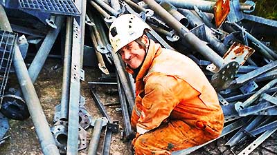 close up of howarths scrap company sheffield manager dressed in the usual high visibilty gear and white hard hat bent down in front of a pile of scrap tubing supports turning his head to the camera and giving a mighty big cheeky looking smile