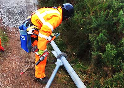 close up of a howarth scrap man outside unbolting two thck metal pipe union joints wearing the usual health and safety bright orange wear with several tool belts around his waste and upper thighs next to a lush green bush