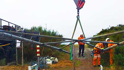 distance center ground outdoor view of a lrafe metal frame suspended from a red crane jib via a thick chain while two howarths experts dessed in high visibility work wear dismantle the corner web joints supervised by a howarths team manager