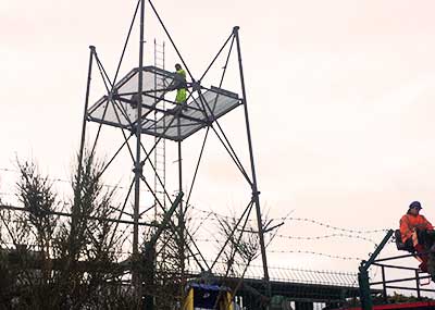 far distance photo of a howarths scrap man wearing bright yellow high visibility jacket and trousers 150 feet up on top of a metal frame mast with tools in hand dismantling an intricate metal framed floor