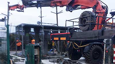 distance view of two howarths scrap men disassembling part of a metal factory side wall with a deep red coloured crane boom overhead complete with a hanging chain ready to attach to the large metal panels